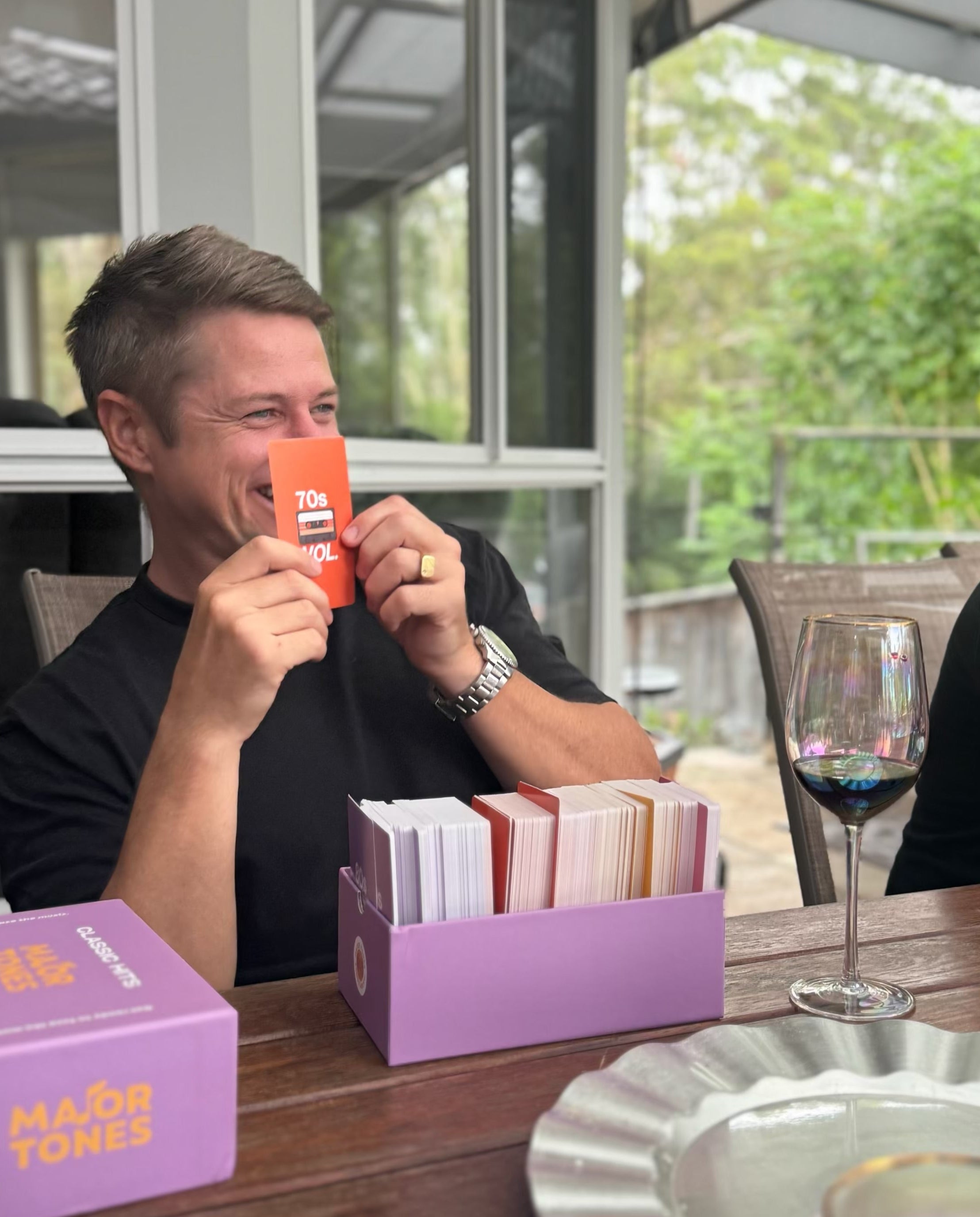 Man in black t-shirt hsitting at table outside holding a 70s card from the Major Tones classic hits deck with the box sitting on the table in front of him